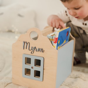 May include: A wooden book holder shaped like a house with a blue door and a window. The house is personalized with the name "Myron". A child is reaching into the book holder to grab a book.