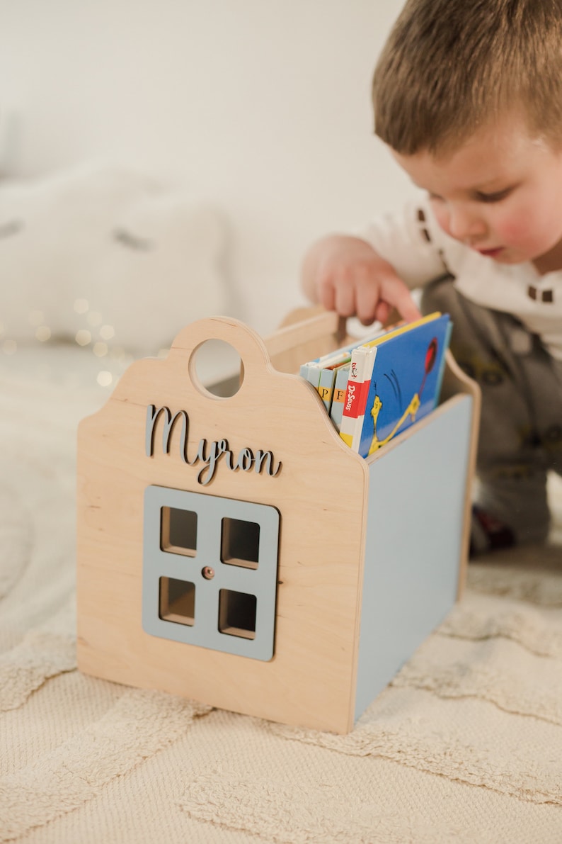 May include: A wooden book holder shaped like a house with a blue door and a window. The house is personalized with the name "Myron". A child is reaching into the book holder to grab a book.