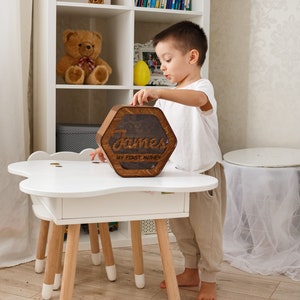 May include: A wooden hexagonal piggy bank with the name "James" and the text "My First Money" engraved on it. A young child is placing the piggy bank on a white table with wooden legs.