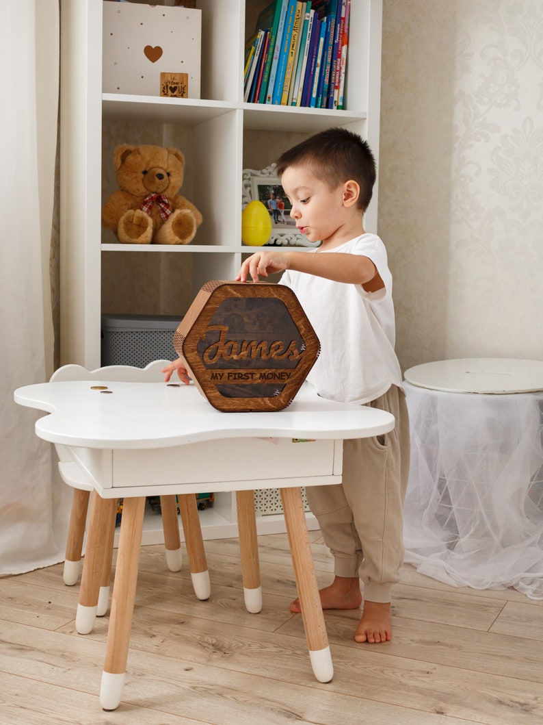 May include: A wooden hexagonal piggy bank with the name "James" and the text "My First Money" engraved on it. A young child is placing the piggy bank on a white table with wooden legs.