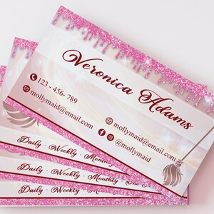 May include: Stack of pink and white business cards with a pink glitter border. The text on the card reads "Veronica Adams" and includes a phone number, email address, and social media handles. The card also features a gold broom with a pink glitter handle.