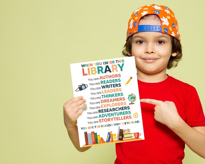 May include: A colorful poster with the text "WHEN YOU ENTER THIS LIBRARY" and a list of roles readers can take on, such as "AUTHORS", "READERS", "WRITERS", "LEADERS", "THINKERS", "DREAMERS", "EXPLORERS", "RESEARCHERS", "ADVENTURERS", and "STORYTELLERS". The poster also includes a small camera, a globe, a magnifying glass, and a pencil. The poster is held by a young person wearing a red shirt and a baseball cap.