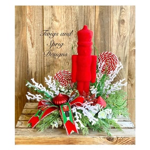 May include: A red velvet nutcracker figurine stands in the center of a Christmas centerpiece. The centerpiece is made of greenery, white berries, and red and white candy canes. The centerpiece is in a wooden crate. The text "Twigs and Sprig Designs" is visible in the image.