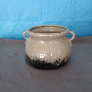 May include: A ceramic pot with two small handles. The pot is a light beige color with a dark brown, brushstroke-like design around the bottom. The background is a solid blue.