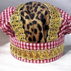 May include: A chef's hat with a red and white checkered pattern, leopard print fabric, and gold trim. The hat is designed with a classic chef's hat shape, featuring a brim and a tall, rounded crown.