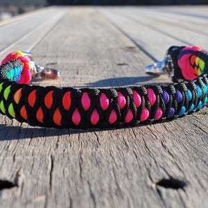 May include: A colorful paracord bracelet with a black base and vibrant rainbow accents. The bracelet features a woven design with pink, orange, yellow, green, blue, and purple details. It has silver-tone clasps.