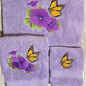 May include: A set of three lavender-colored bath towels. Each towel features an embroidered design of a monarch butterfly with orange and black wings, purple flowers, and green leaves. The towels are made of a soft, absorbent material.