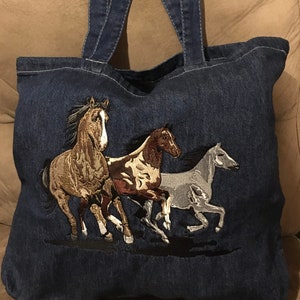 May include: A blue denim tote bag with three embroidered horses running. The horses are brown, white, and gray.