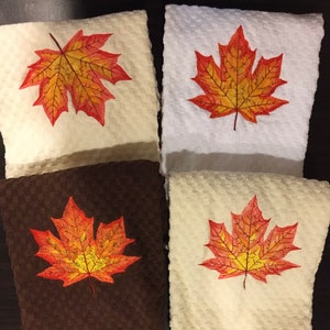 Puede incluir: Cuatro toallas de cocina con hojas de arce naranja y amarillo bordadas. Las toallas son de varios colores, incluyendo blanco, marrón y beige.