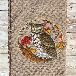 May include: A brown kitchen towel with an embroidered design of an owl perched on a branch with autumn leaves. The owl has yellow eyes and brown and white feathers.