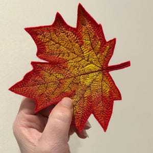 Puede incluir: Hoja de arce bordada en tonos rojos, naranjas y amarillos. La hoja presenta costuras detalladas que resaltan las venas y los bordes. La hoja se sostiene sobre un fondo neutro.