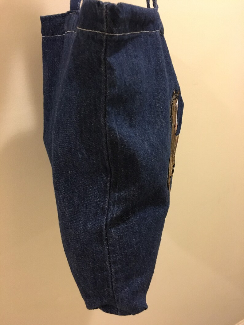 May include: A blue denim tote bag with a gold embroidered design on the side.