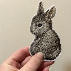Puede incluir: Parche bordado que representa un conejo en tonos marrones y grises. El conejo se muestra de perfil, con las orejas levantadas y un cuerpo esponjoso. El parche tiene un borde blanco.