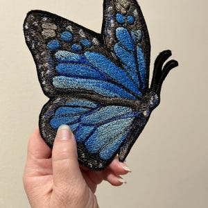 May include: Embroidered butterfly patch featuring shades of blue and black. The butterfly's wings showcase intricate details with a gradient of blue hues, and a black border. This decorative patch is ideal for adding a unique touch to clothing or accessories.