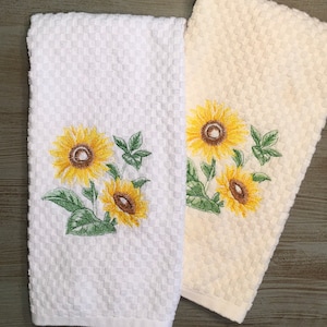 May include: Two kitchen towels with embroidered sunflowers. One towel is white and the other is a light cream color.