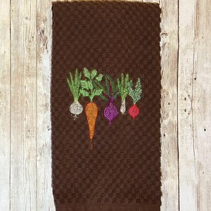 May include: Brown kitchen towel with an embroidered design of five vegetables: a green onion, a carrot, a beet, a turnip, and a radish.