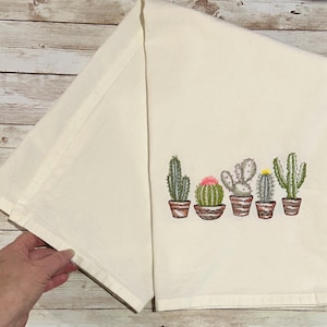 May include: Cream-coloured kitchen towel featuring an embroidered design of five potted cacti. The cacti are green, grey, and pink, in brown pots. The towel is folded and held by a hand.