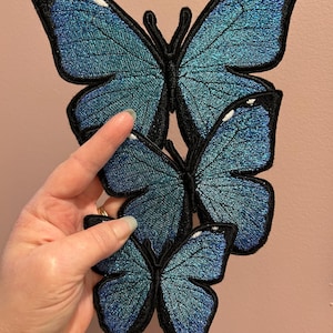 May include: Three blue butterfly embroidered patches with black outlines. The butterflies are layered on top of each other, with the largest butterfly on the bottom and the smallest butterfly on top.
