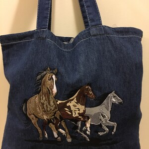 May include: A blue denim tote bag with three embroidered horses running. The horses are brown, tan, and gray.