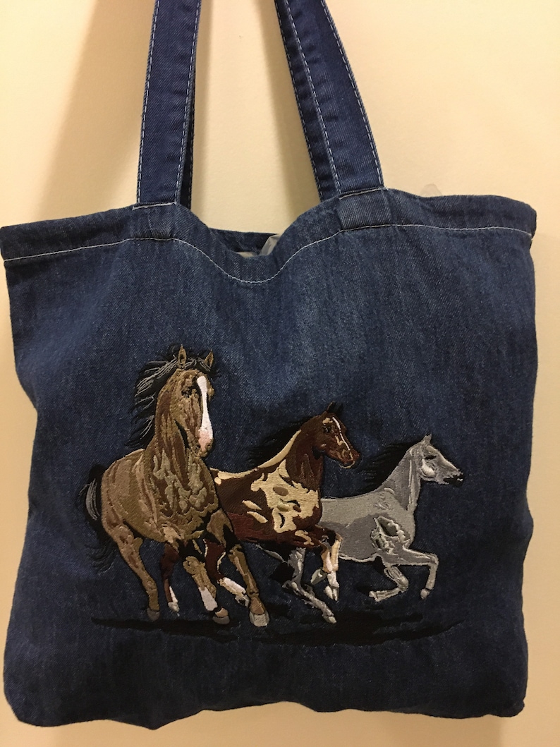 May include: A blue denim tote bag with three embroidered horses running. The horses are brown, tan, and gray.