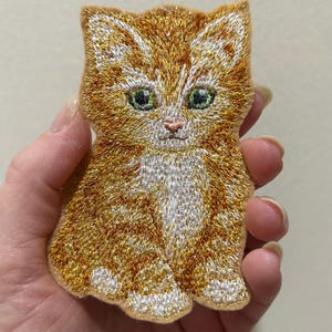 May include: Embroidered patch of a ginger kitten with green eyes and white chest. The patch is made with orange, gold, and white thread, and is in the shape of a sitting cat. The patch is likely for clothing or accessories.