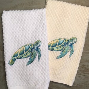 May include: Two kitchen towels with an embroidered sea turtle design. One towel is white and the other is a light beige color.