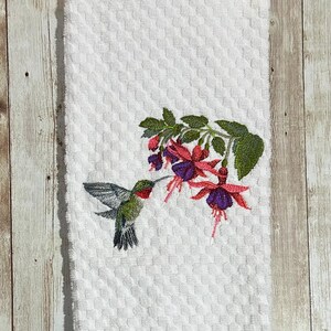 May include: White hand towel with a textured pattern. An embroidered hummingbird is shown feeding from a fuchsia flower. The hummingbird has green, gray, and red coloring. The fuchsia flowers are pink and purple.