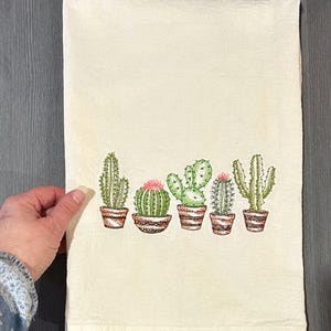 May include: Cream-coloured kitchen towel with an embroidered design of five potted cacti. The cacti are green with pink flowers, in brown pots. The towel is folded and set against a grey background.