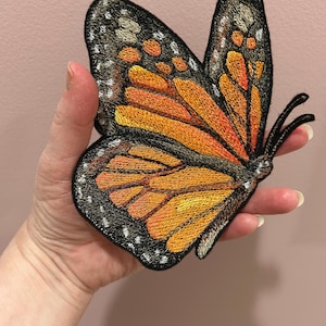 Puede incluir: Parche de mariposa bordado con detalles en naranja, negro y blanco. El parche presenta un diseño detallado de una mariposa monarca, con intrincados patrones de alas y un borde negro. El parche de mariposa se sostiene en una mano.