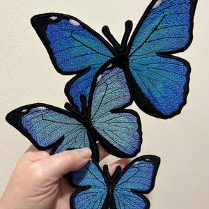 Puede incluir: Tres parches bordados de mariposas de diferentes tamaños. Las mariposas presentan tonos de azul con contornos y antenas negros. Los parches se sostienen sobre un fondo neutro, mostrando las costuras detalladas y los degradados de color.