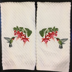 May include: Two white kitchen towels with embroidered hummingbirds and fuchsia flowers. The hummingbirds are green, black, and white. The flowers are pink and red.