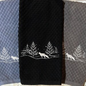 May include: Three kitchen towels with a winter scene embroidered on them. The towels are blue, black, and gray. The scene features a fox walking through a snowy forest with trees.