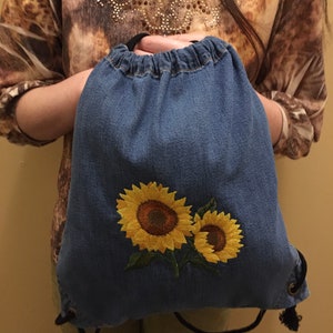 Peut inclure: Un sac à cordon en jean bleu avec deux tournesols brodés. Les tournesols sont jaunes avec des centres bruns et des feuilles vertes.