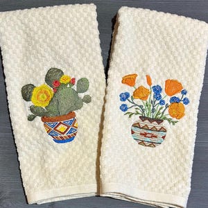 May include: Two off-white, textured hand towels with embroidered designs. One features a cactus with yellow flowers in a patterned pot. The other has orange and blue flowers in a similar pot. The towels have a woven pattern.