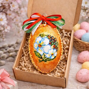 May include: An Easter egg ornament in a cardboard box, decorated with a floral design. The egg is orange with a painted image of blue and white flowers. A red and green ribbon is tied at the top. Decorative Easter eggs are in the background.