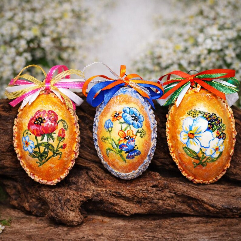 May include: Three decorative, hand-painted Easter eggs with floral designs. Each egg features a different flower arrangement in shades of red, blue, and white, with colorful ribbon bows. The eggs are set against a blurred, natural background.