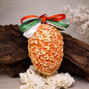 May include: An orange, textured, egg-shaped ornament with gold accents, topped with a bow of red, green, and white ribbons. The ornament is displayed on a piece of white coral and a piece of driftwood.