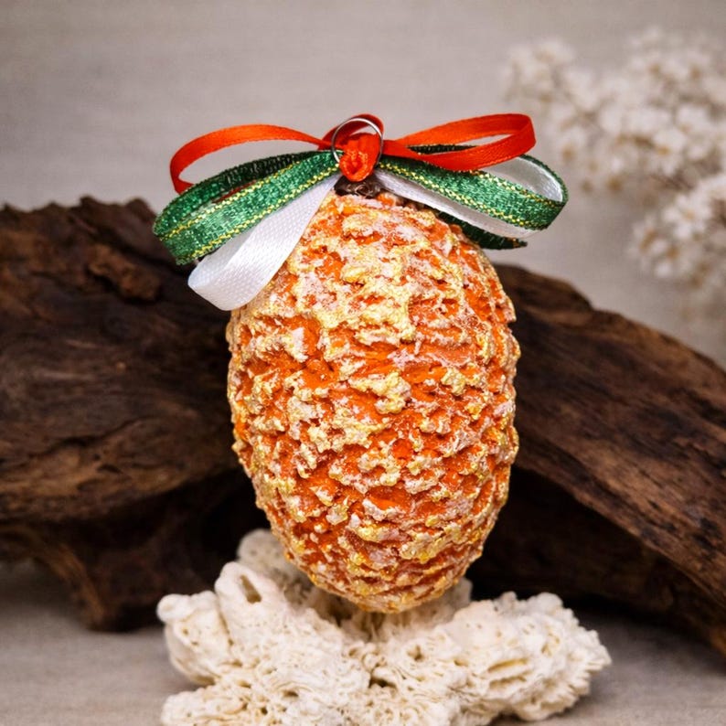 May include: An orange, textured, egg-shaped ornament with gold accents, topped with a bow of red, green, and white ribbons. The ornament is displayed on a piece of white coral and a piece of driftwood.