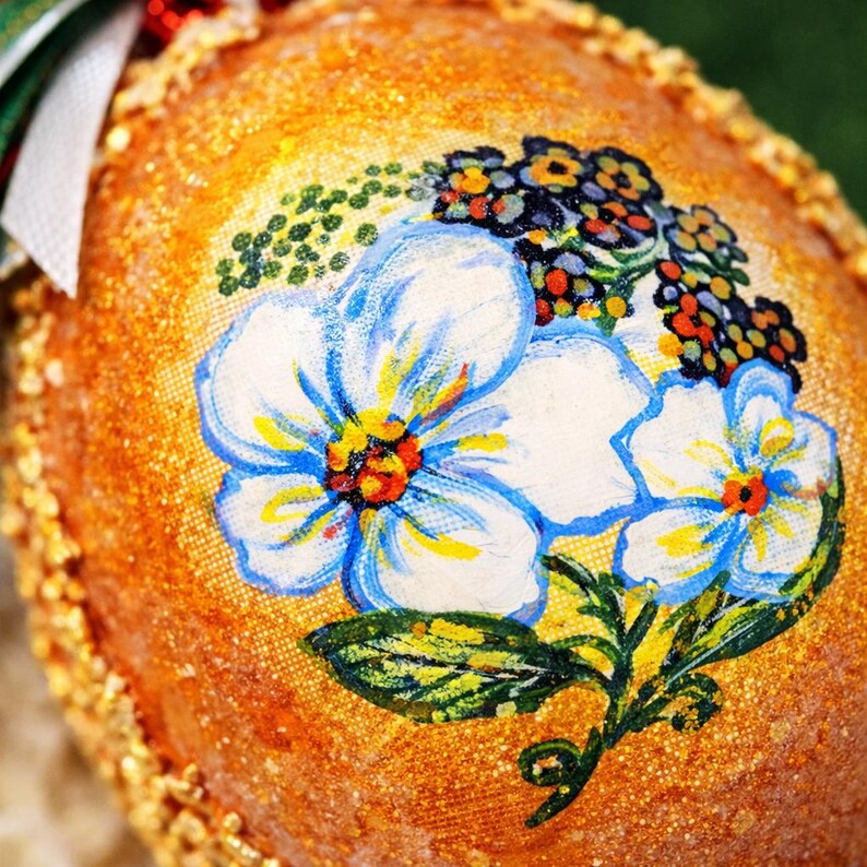 May include: Close-up of a decorative, egg-shaped ornament. The ornament has a textured, golden-orange surface with a floral design. The design features white flowers with blue outlines and yellow centers, along with green leaves and small berries. A green and white ribbon is visible.