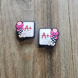 May include: Two pink and black pencil charm shoe charms with a white paper with the text "A+" and a white flower.