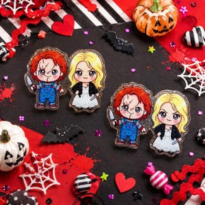 May include: Two pairs of acrylic pins featuring chibi-style characters with red hair in blue overalls and blonde hair in white dresses with black jackets. Each character holds a small knife. The pins have a glittery finish and clear edges.