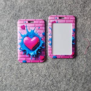 May include: Two pink ID badge holders with a heart design. One holder features a pink heart on a blue splatter against a pink brick wall background. The other holder is empty with a clear window.