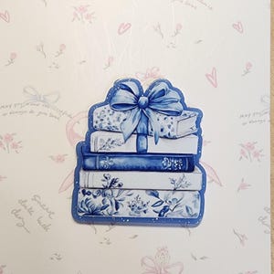 May include: A blue and white stack of books with a blue bow on top. The books have floral and patterned designs. The background is a light pink with floral and heart designs and the text "May you have the base..." and "Sweet duke luck day."