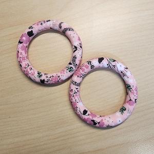 May include: Two pink floral print bangle bracelets. The bracelets have a light pink base color with a repeating pattern of pink flowers, black accents, and green leaves. The bracelets are circular in shape.
