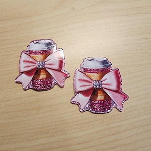 May include: Two pink glitter coffee cup charms with a pink bow. The coffee cups have a white lid and a brown liquid inside.
