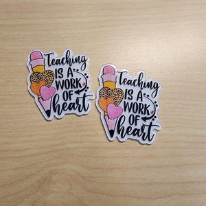 May include: Two clear stickers with a pencil design and the text "Teaching is a work of heart." The pencil has a pink eraser, a leopard print section, and a pink heart. The stickers are on a light wood surface.