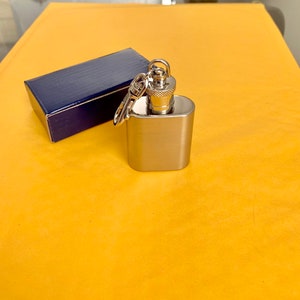 May include: A small, stainless steel flask with a screw-on cap and attached keychain. The flask is rectangular with rounded edges and sits next to a small, dark blue box. The background is a solid yellow color.