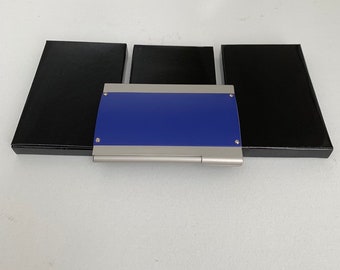 May include: A blue and silver business card holder with three black boxes. The card holder has a rectangular blue top with silver trim. The boxes are rectangular and black. The items are arranged on a white surface.