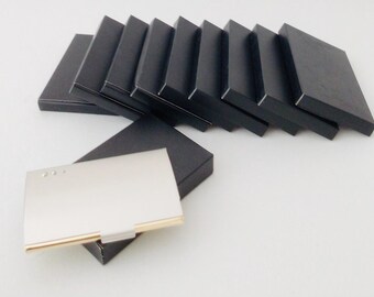 May include: A silver business card holder with gold trim is displayed with several black boxes. The card holder has a rectangular shape with three small decorative accents. The black boxes are arranged in a row, suggesting a set or collection.