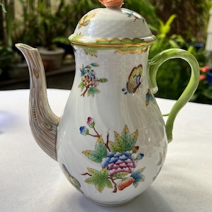 May include: A white porcelain teapot with a floral and butterfly design. The teapot has a green handle, a brown spout, and a rose-shaped knob on the lid. The floral design includes blue, pink, and yellow flowers.